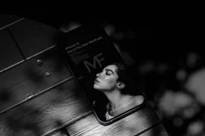 A smartphone displaying a black and white photo of a woman's face, resting on a wooden surface in dim lighting, serves as an ideal mockup. - PSD Mockup