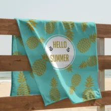 A turquoise beach towel with a pineapple pattern and the template "hello summer" draped over a wooden railing with a sandy beach and ocean in the background. - PSD Mockup