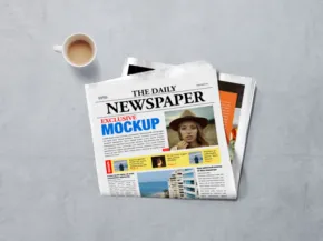 A newspaper template lies next to a coffee cup on a textured gray surface, featuring articles and images. - PSD Mockup