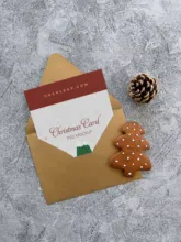 A Christmas card template peeks out from an envelope next to a gingerbread man and pine cone on a textured surface. - PSD Mockup