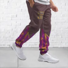 Man wearing brown sports pants with template purple and gold accents and white sneakers standing against a mockup white brick wall. - PSD Mockup