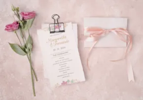 Wedding invitation template on a clipboard with a pink ribbon, flowers, and an envelope on a textured background. - PSD Mockup