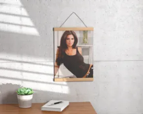 Mockup of a woman in a black top, framed and hanging on a sunlit wall above a small table with a plant and a book. - PSD Mockup
