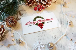 A "merry christmas" mockup card with a floral wreath design, surrounded by pine cones, berries, and string lights on a wooden surface. - PSD Mockup