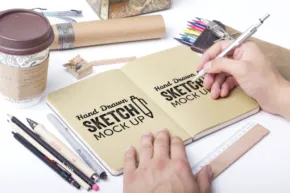 A person sketches in a notebook labeled "sketch book," surrounded by art supplies and a coffee cup. - PSD Mockup