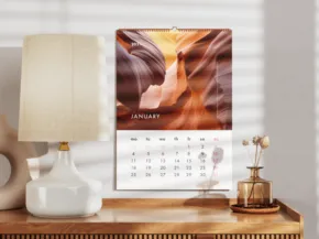 A calendar with a landscape image hangs on a wall beside a lamp and decorative objects on a white desk. Sunlight filters through blinds, casting shadows. - PSD Mockup