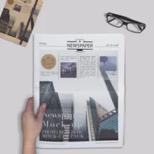 A person holding a newspaper with articles and images, next to a pair of eyeglasses on a table, serving as a template. - PSD Mockup