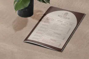 A mockup of a restaurant menu on a wooden table next to a potted plant. - PSD Mockup