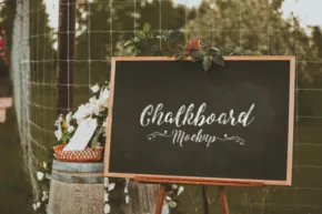 A rustic wedding sign template reading "chalkboard our wedding day" on a blackboard, adorned with white flowers and greenery, set outdoors. - PSD Mockup