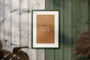 A framed mockup with the words "exclusive frame work" on a green wooden background, partially obscured by plant leaves in the foreground. - PSD Mockup