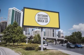 A large billboard with the template "outdoor billboard mockup" stands beside a road in an urban area with skyscrapers and a clear blue sky. - PSD Mockup