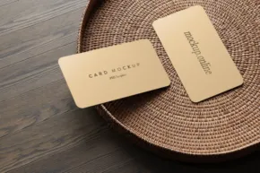 Two cream-colored business cards with gold embossing, presented as a mockup on a textured straw hat on a wooden surface. - PSD Mockup
