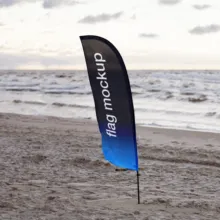 A vertical banner with the text "flag mockup" displayed on a beach at sunset, with waves in the background. - PSD Mockup