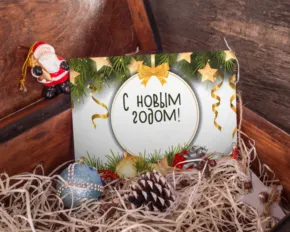 Holiday greeting card template with "с новым годом!" text, surrounded by Christmas decorations, pine cones, and a Santa figurine inside a wooden box. - PSD Mockup