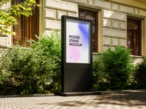 A digital advertising mockup stands on a sidewalk, featuring a colorful gradient template, surrounded by lush green bushes outside a classic building. - PSD Mockup