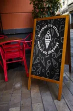 A chalkboard mockup advertising ice cream, placed on a sidewalk next to a red chair and a large planter, with various ice cream illustrations. - PSD Mockup