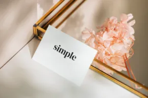 A white card template with the word "simple" printed on it, resting on a surface alongside a pink crystal and golden thin rods, with soft sunlight casting shadows. - PSD Mockup