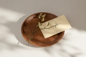 A wooden bowl containing a small branch and a card with handwritten text, serving as a mockup, casting a soft shadow in sunlight. - PSD Mockup