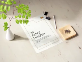 A4 paper template on a desk with a white ceramic vase holding green leaves and ink pads nearby. - PSD Mockup