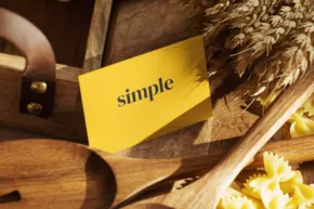 A close-up photo of dry pasta shapes on a wooden spoon, with wheat and a yellow mockup labeled "simple" in the background. - PSD Mockup