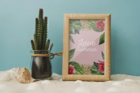 A decorative setting with a cactus in a glass vase, a framed "have a nice summer" template, and a seashell on a sandy surface against a light blue background. - PSD Mockup