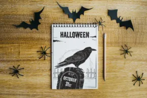 Notebook template with "halloween" written and a crow drawing on a wooden surface, surrounded by paper bats and spider decorations. - PSD Mockup