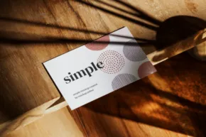 A business card template with the word "simple" printed on it, resting on a wooden surface with sunlight casting diagonal shadows. - PSD Mockup