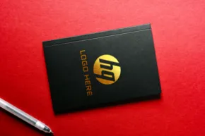 A black business card template with a gold hp logo lying on a red surface next to a silver pen. - PSD Mockup