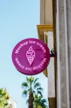 Purple mockup sign reading "make-up ice cream, looks and dessert" hanging on a building, with a palm tree and the sky visible in the background. - PSD Mockup