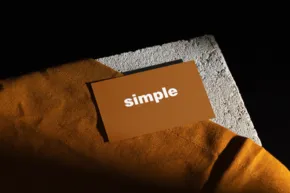 A card with the word "simple" rests on a textured orange blanket beside a gray edge, against a dark background designed as a mockup template. - PSD Mockup