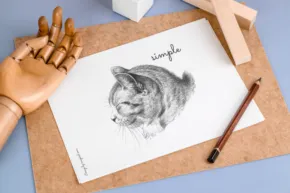 A pencil sketch of a cat on paper, signed at the bottom, with a wooden hand model and a pencil resting beside it on a blue background. - PSD Mockup