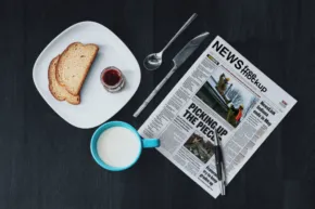 A breakfast template featuring a cup of milk, toast with jam, and an open newspaper on a dark table. - PSD Mockup