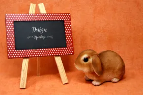 A small brown rabbit sits beside a miniature blackboard with "design got me like" written in chalk, set against an orange background, serving as a perfect mockup. - PSD Mockup
