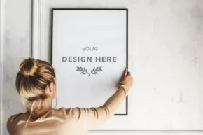 Person attaching a framed poster template with the text "your design here" to a white wall. - PSD Mockup