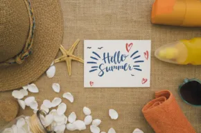 A flat lay template of summer beach essentials with a 'hello summer' card, straw hat, sunglasses, sunscreen, sandals, towel, and seashells on a sandy background. - PSD Mockup