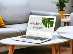 A laptop open on a round white table in a cozy room, displaying the words "macbook free template" on its screen, with a sofa and decorative items in the background. - PSD Mockup