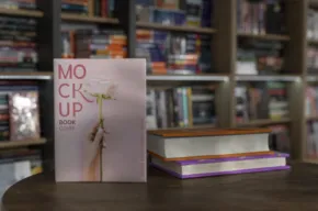 A book with "mockup" on the cover stands upright on a wooden surface, with a blurred bookshelf in the background. - PSD Mockup