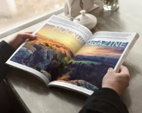 Hands holding an open magazine with a mockup of a landscape photo on the page, by a window with natural light. - PSD Mockup