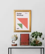 A framed poster template on a white wall above a console table with a globe, books, and a potted plant. - PSD Mockup