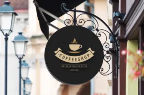 Round black sign template with a coffee cup logo and the text "coffeehouse" hanging from a wrought iron bracket on a building facade. - PSD Mockup