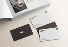 Two business cards featuring a minimalist design with a white and dark brown color scheme, resting beside an open brochure on a light surface, serve as a perfect mockup template. - PSD Mockup