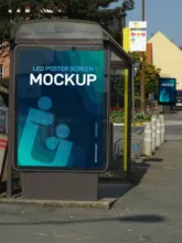 A bus stop shelter featuring a template poster screen with a mockup advertisement, located on a sunny street with trees and a building in the background. - PSD Mockup