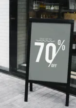 A mockup of a sidewalk sign outside a store displaying a "70% off" advertisement in bold white text on a black background. - PSD Mockup