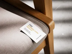 Close-up of a product tag mockup on a tan upholstered chair with a wooden frame, placed under soft lighting with striped shadows in the background. - PSD Mockup