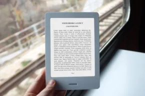 A person holding an e-reader mockup with text on the screen by a window with a blurred background, suggestive of a train journey. - PSD Mockup
