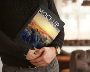 Person holding a magazine with "mockup" written on the cover, featuring a landscape image, in a cozy indoor setting. - PSD Mockup