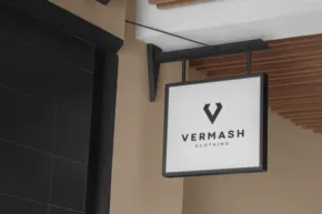 A hanging rectangular black and white store sign mockup with the logo "v" and the word "vermash" displayed on a building facade. - PSD Mockup
