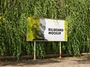 A billboard template with a partial green overgrowth standing in front of dense foliage in a park setting. - PSD Mockup