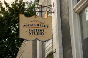 A mockup of a hanging sign for "master line tattoo studio" mounted on a building's exterior wall, with trees and sky in the background. - PSD Mockup