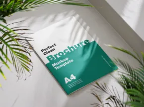 A brochure labeled "business brochure mockup" lays on a white surface, partially shadowed by palm leaves, with a sunlight accent. - PSD Mockup